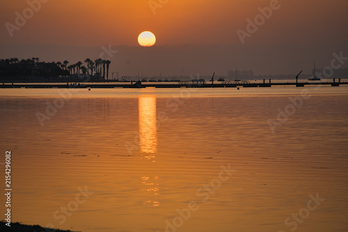 Golden sun dips low over calm waters. Silhouetted palm trees and distant structures frame the horizon. Ripples reflect the sun's warm glow across the surface. Misty atmosphere adds serene