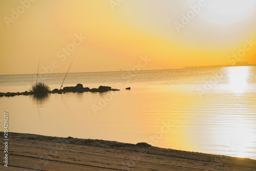 Golden sun dips low, casting warm glow. Ripples dance across calm, reflective water.  Beach sand stretches gently toward the sea