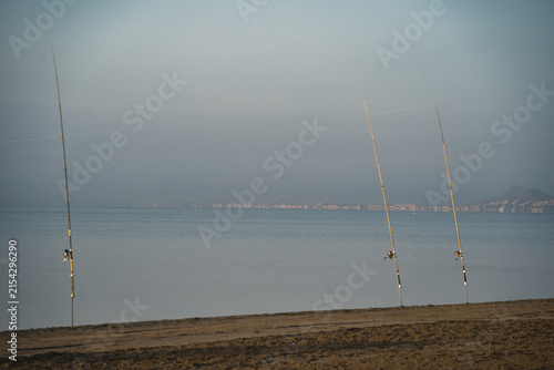 Three fishing rods stand upright on sandy beach. Still waters stretch to distant, hazy shoreline. Gray sky suggests early morning or late dusk. Empty scene evokes quiet solitude and patience