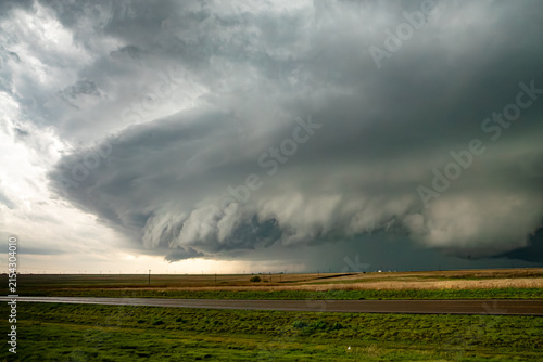 View of a massive supercell storm cloud looming over a flat rural landscape with a road in the foreground.