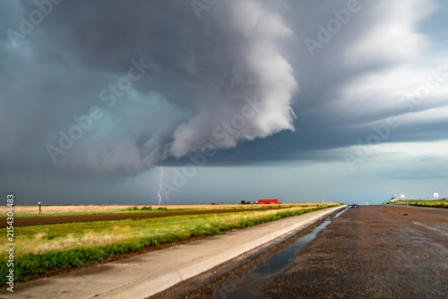 View of a supercell storm with a lightning bolt striking over a rural landscape with green fields and a distant red building under dark, ominous clouds.