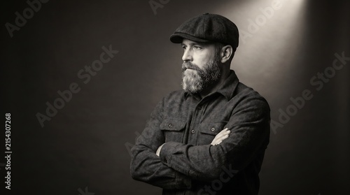 Portrait of a bearded man in a flat cap and shirt standing with arms crossed in studio.