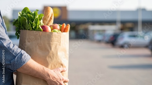 Person stands in a parking lot holding a paper bag filled with fresh produce and baked goods. The supermarket is in the background under the afternoon sun