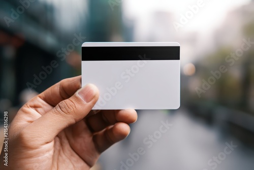 Close up of male hand holding blank white credit card mockup with black magnetic stripe. Blurred city street background. Commercial payment template for bank debit plastic, urban discount voucher