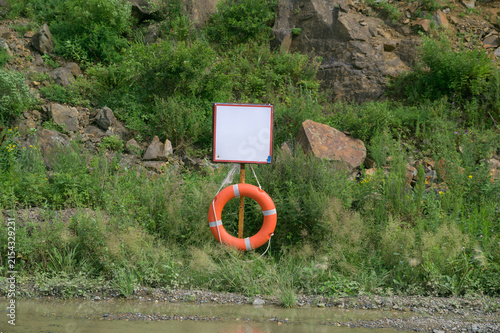 Orange lifebuoy beneath blank sign on rocky green shore.
