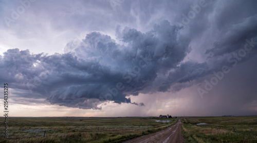 Powerful supercell thunderstorm cloud formation over a vast rural landscape with a dirt road and distant farmhouse.