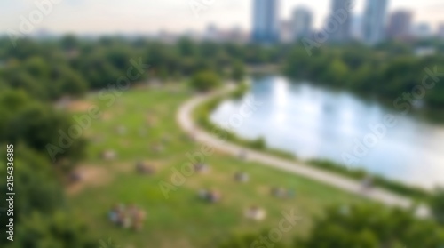 Aerial view of Central Park lake and green lawn with people relaxing under bright sky and blurred city skyline in distance during warm summer day in New York City
