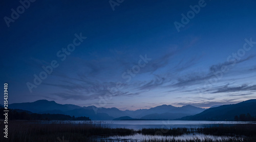 Peaceful blue hour lake panorama framed by majestic mountains and delicate reeds under a vast blue sky.