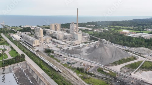 High-angle aerial shot circling the large concrete silos, kiln exhaust stack, raw material aggregate piles, and freight train tracks of a cement factory on Lake Ontario in Mississauga, Canada