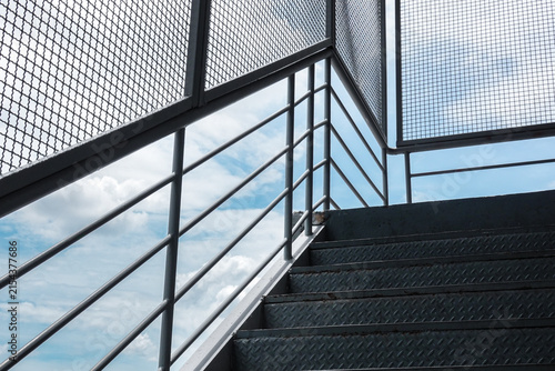 Low angle view of modern industrial metal staircase with wire mesh railing against blue sky clouds.