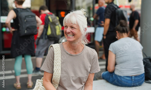 Happy senior woman laughing while waiting at bus station in the city - Travel, lifestyle and mature person concept