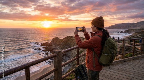 Woman photographing scenic ocean sunset from viewpoint, capturing coastal landscape with smartphone, travel photography inspiration