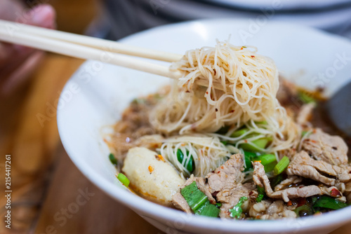 Chopsticks lifting thin rice noodles from Thai boat noodle soup with pork and meatballs in white bowl.