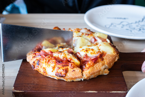 Stainless steel spatula lifting a slice of Hawaiian pizza with pineapple ham and melted cheese on wooden board.