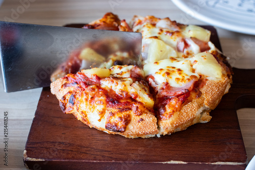 Stainless steel spatula lifting a slice of Hawaiian pizza with pineapple ham and melted cheese on wooden board.