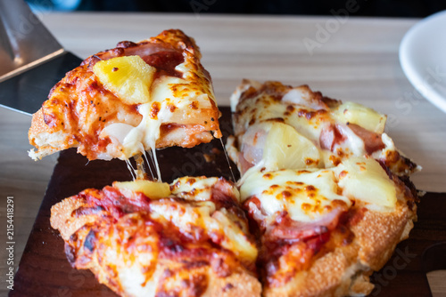 Lifting a slice of Hawaiian pizza with melted cheese pull and pineapple toppings using metal spatula.