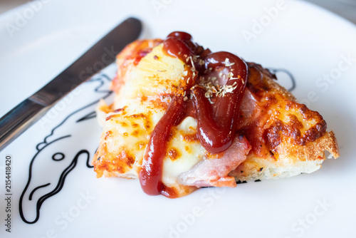 Hawaiian pizza slice drizzled with tomato ketchup and oregano served on white plate with fork and knife.