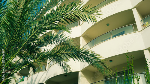 Modern hotel building facade with balconies behind lush green palm trees. Summer vacation resort concept, tropical holiday accommodation.	
