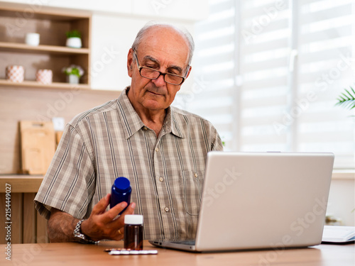 Senior man with eyeglasses using laptop for searching details about his prescribed medication while sitting at his cozy home. Telemedicine concept.