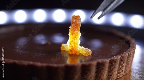 Hand using tweezers to place vibrant orange candy decoration on a chocolate tart, showcasing meticulous attention to detail in a well-lit kitchen setting