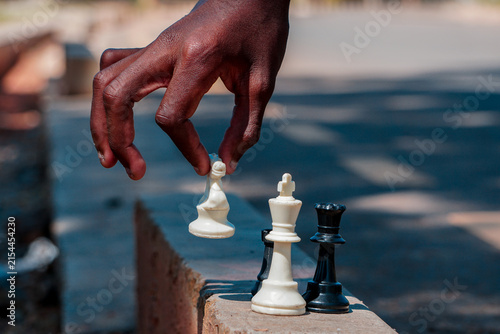 Obraz na płótnie A close-up of a dark-skinned hand moving a white chess piece on an outdoor stone ledge, alongside a white king and black queen in bright sunlight