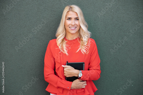 Blonde professional woman smiling, holding a notebook, standing confidently against a green wall, representing business success