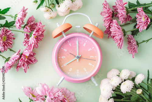 Pink alarm clock surrounded by blooming flowers on a soft green background, decorative flat lay. Top view