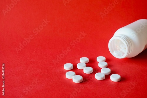 Konstfotografi White round medical pills scattered on a solid red surface next to an open white plastic bottle
