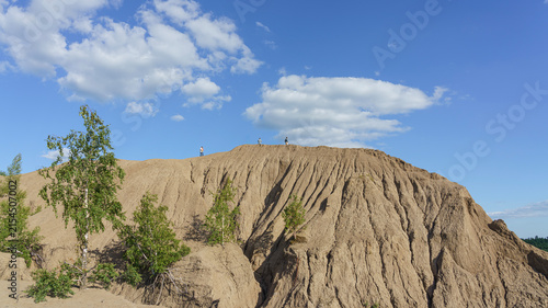 Obraz na płótnie Unrecognizable people standing on a clay hilltop with distinct ridges under a blue sky with scattered clouds, surrounded by sparse vegetation and trees in Konduki, Tula region, Russia