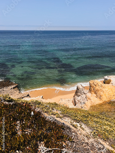 Scenic coastal view of a small sandy beach surrounded by rocky cliffs and coastal vegetation, with clear turquoise ocean water stretching to the horizon