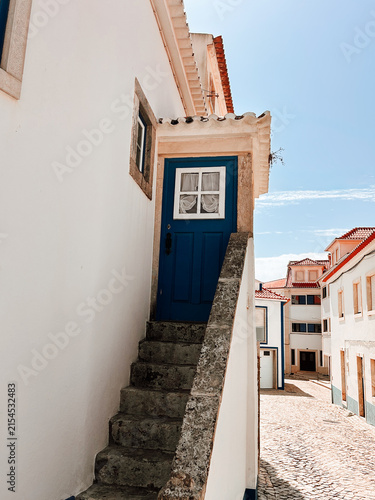 Traditional blue wooden door reached by a narrow exterior stone staircase on a whitewashed house, with tiled roofs and cobblestone street in the background