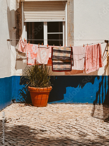 Laundry hangs on a clothesline outside a window above a blue and white wall, with a terracotta plant pot and cobblestone pavement in bright sunlight