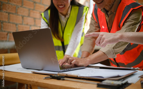 Team of Asian engineers collaborating on blueprints and a laptop inside a construction site.