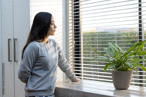 Woman in grey sweater standing at home window gazing through blinds with potted plant on windowsill