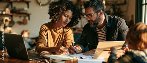 Parent dining table family planning laptop notebook home kitchen child adult discussion with paper, pen, warm light, mixed ethnicity couple, calm expression, domestic routine