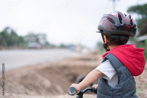 Young boy wearing a helmet and red-hooded jacket, holding a balance bike and looking at a construction area by the road, captured from behind.