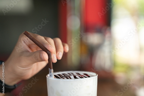 Close-up of a hand inserting a brown straw into a glass of iced milk topped with chocolate syrup, with a blurred café background and soft natural light.