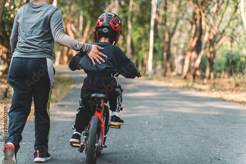 Parent supporting child learning to ride a red bicycle in a park, captured from behind on a shaded path through the woods.