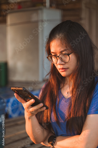 Young woman with glasses focused on her smartphone in warm sunlight.