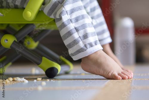 Young child's bare feet dangling from a high chair with messy food leftovers on the floor.