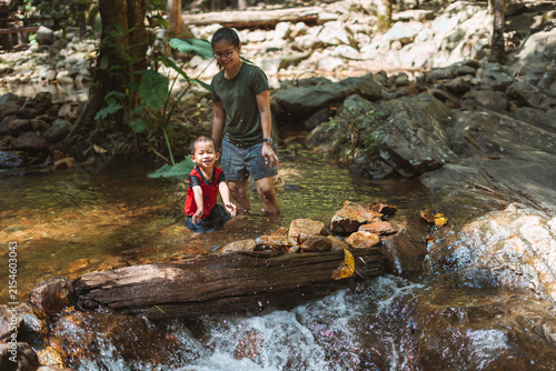 Mother and young boy playing and enjoying a shallow forest stream near a small waterfall surrounded by rocks and greenery.