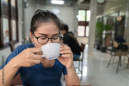 Young woman with glasses enjoying a warm beverage from a white cup in a cozy modern cafe, surrounded by a calm indoor atmosphere.