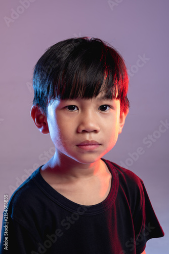 A young Thai boy with a serious expression is illuminated by dramatic neon lighting in a studio portrait.