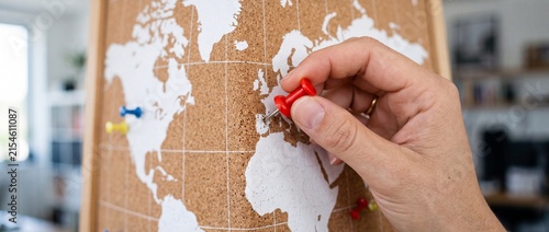Hand placing red push pin on paper world map attached to cork board surface for travel planning global logistics navigation business goals