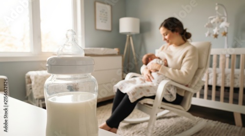 A milk bottle in focus with a mother nursing an infant in a blurred nursery background.