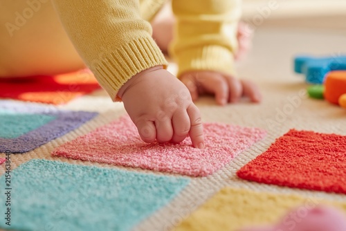 Close up of toddler's hand exploring colorful textured fabric squares on a soft play mat, with additional toys visible in the background