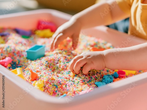 Close up of toddler hands exploring colorful sensory play materials in a pink container filled with vibrant grains and small toys, bright indoor setting