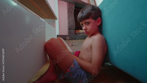 Angry boy hiding behind sofa at home, withdrawn stare expressing frustration, misunderstood childhood trauma, emotional distress and deep social isolation