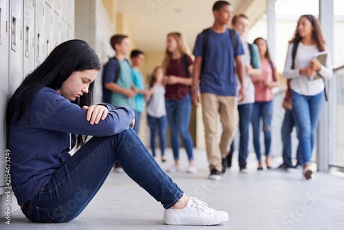 A sad and lonely teenage girl sitting on the floor in a school hallway leaning against lockers while other students walk past in the background.