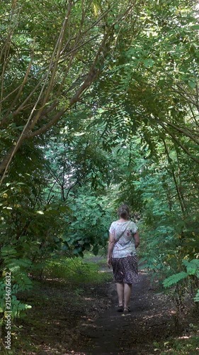Rear view of a woman strolling deeper along a narrow woodland trail under an arch of leafy branches in soft summer daylight